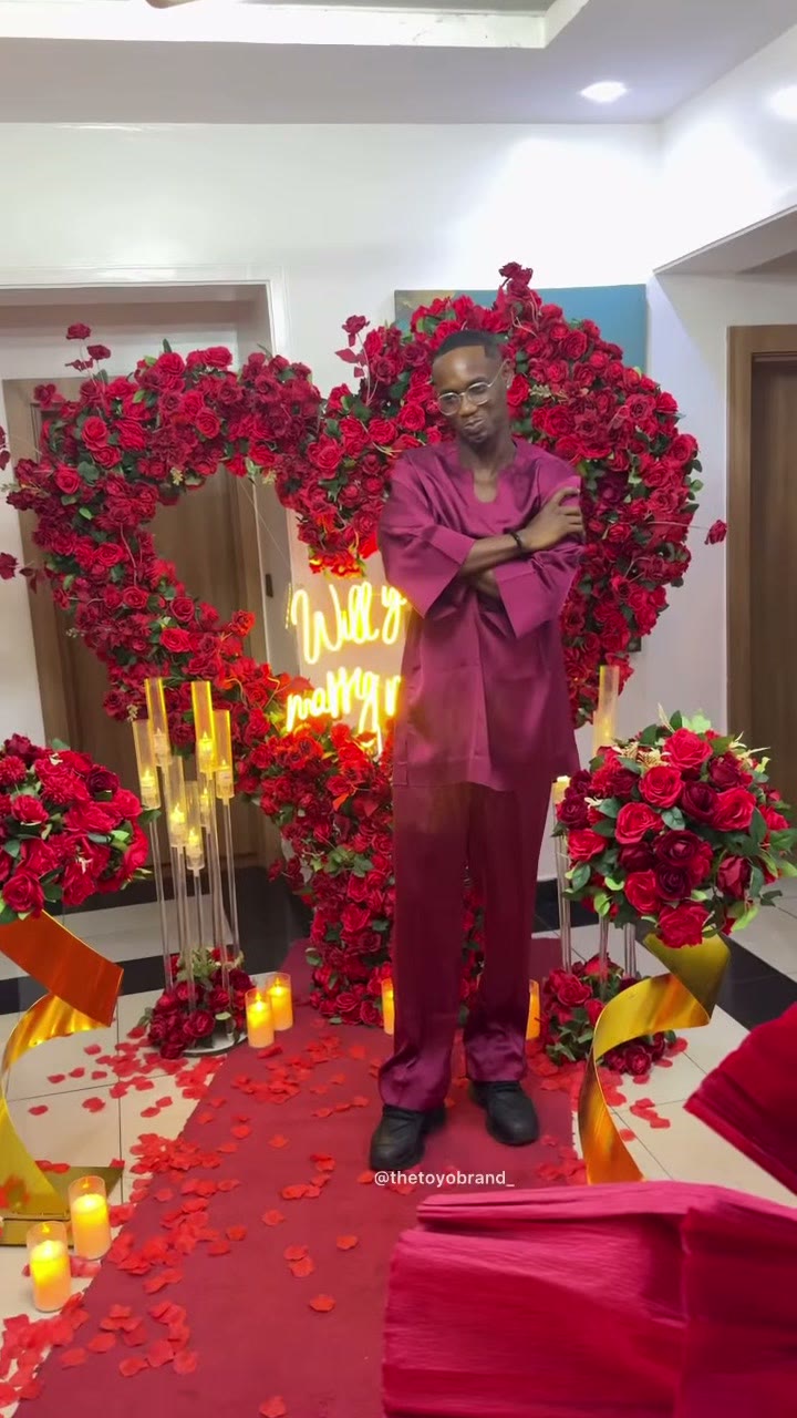 A heart-shaped red-rose arch with a gold neon will-you-marry-me sign and a candlelit aisle